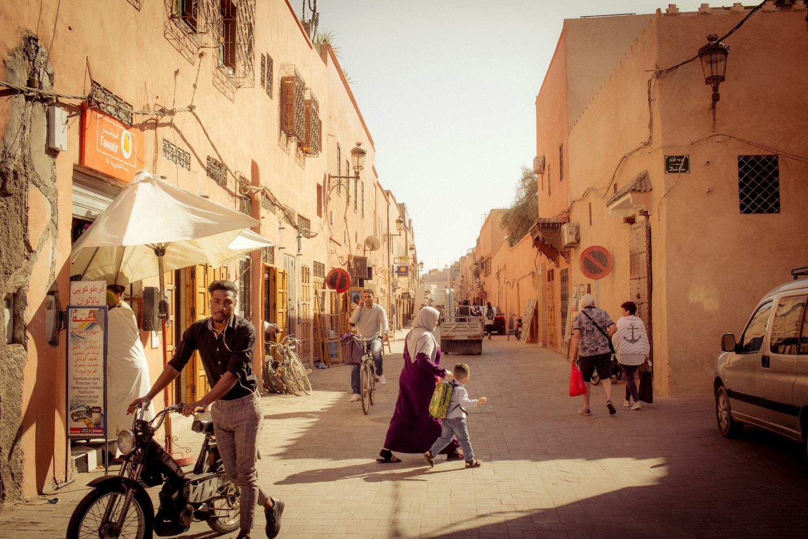 أشخاص يسيرون في شارع ضيق بين مبانٍ سكنية عالية وقت العصر